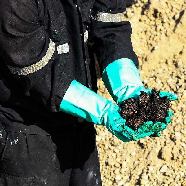 A person in black protective clothing and rubber gloves holding black oily material