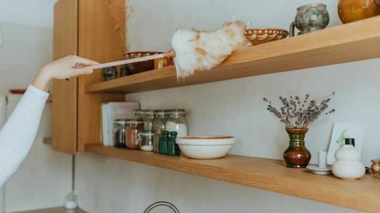 A person running a duster over a tall shelf in a kitchen