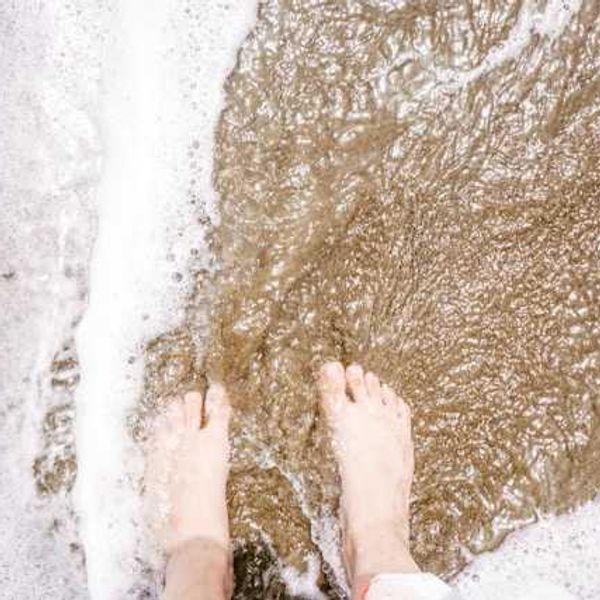 A person's feet on the beach with foamy surf coming in