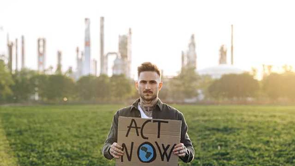 A person standing in front of a power plant holding a sign saying Act Now
