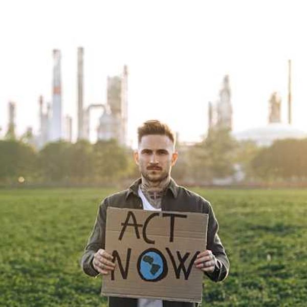 A person standing in front of a power plant holding a sign saying Act Now