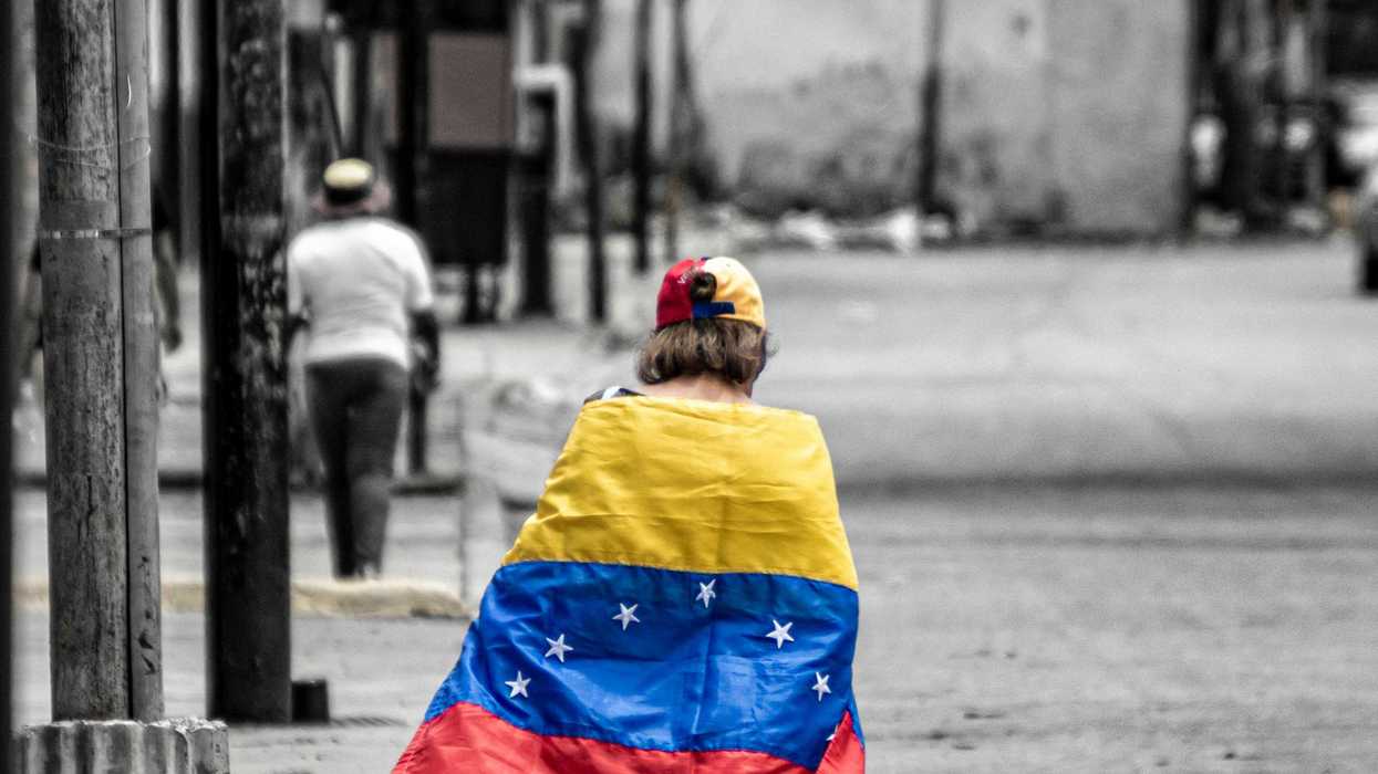 A person walking down a drab gray street wrapped in a colorful Venezuelan flag.