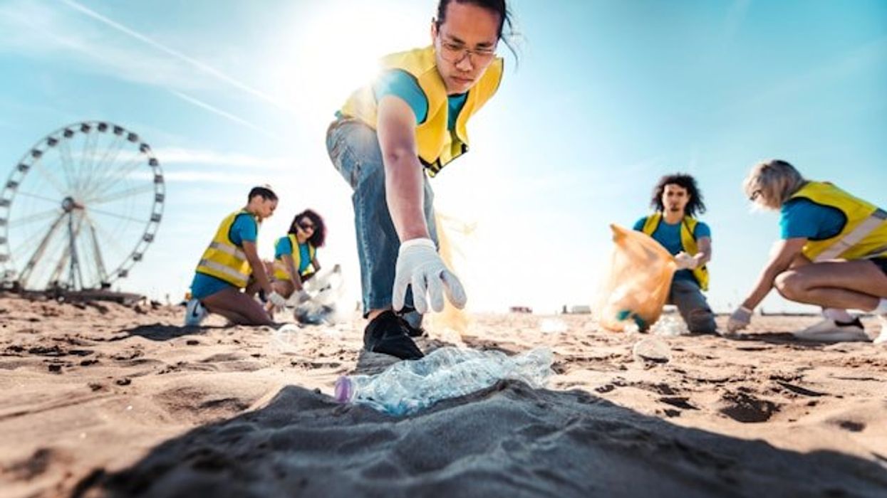A person wearing a yellow safety vest reaching for a plastic bottle on the beach