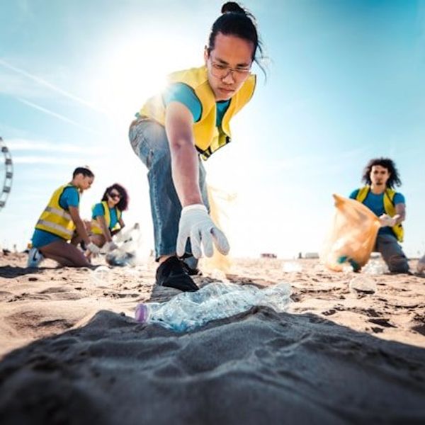 A person wearing a yellow safety vest reaching for a plastic bottle on the beach