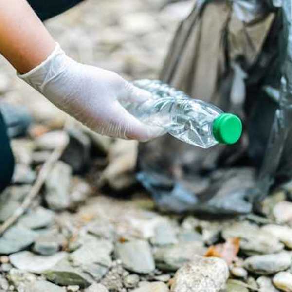 A person wearing plastic gloves picks up a plastic bottle from a rocky beach