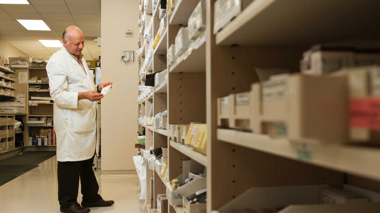 A pharmacist in a white coat examines a package of medication in front of a shelf stocked with prescription medicines.