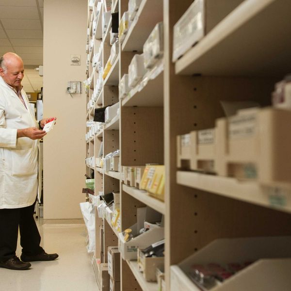 A pharmacist in a white coat examines a package of medication in front of a shelf stocked with prescription medicines.