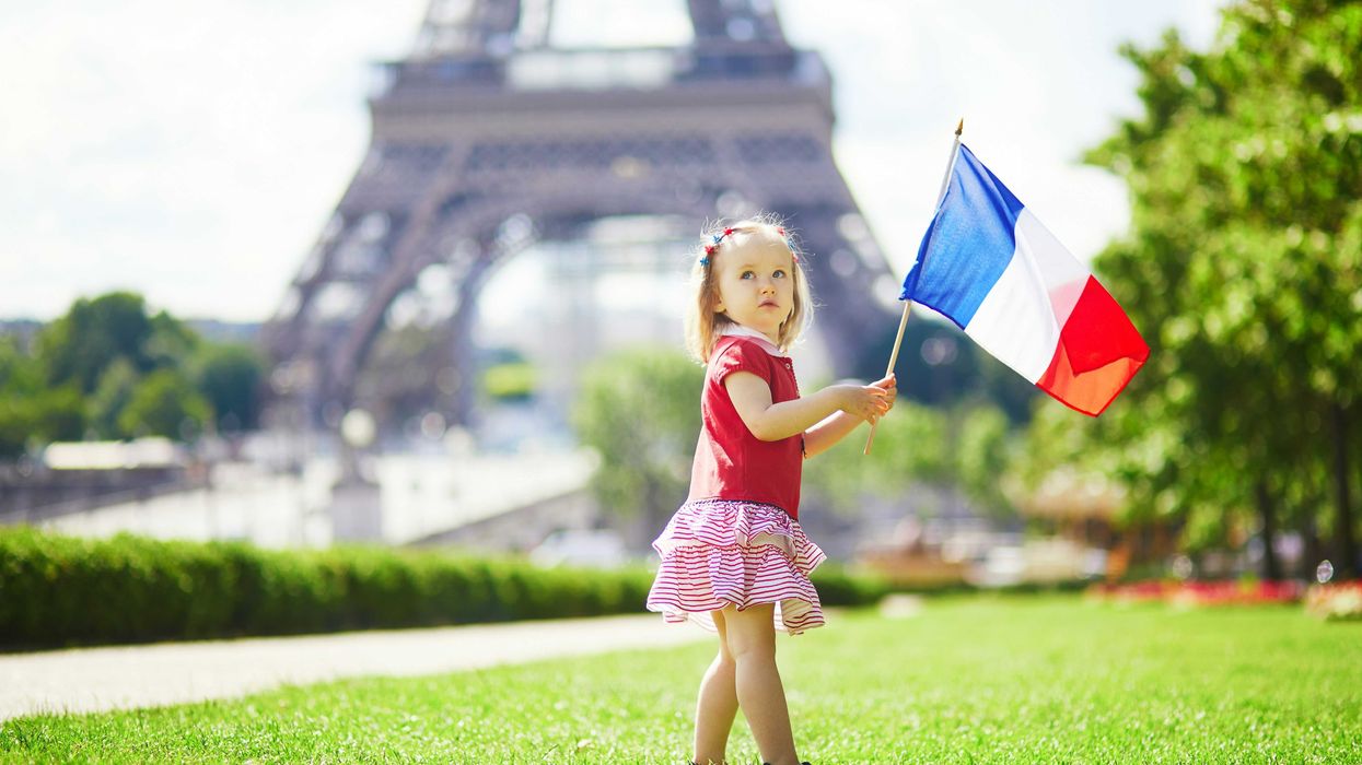 A pre-school aged girl waves the French flag on a lawn with the base of the Eiffel Tower in background.