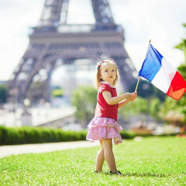 A pre-school aged girl waves the French flag on a lawn with the base of the Eiffel Tower in background.