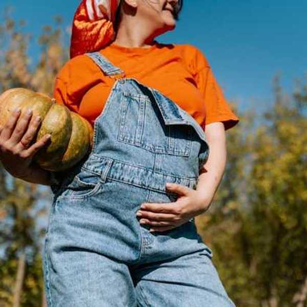 A pregnant woman wearing overalls and holding her belly, with a squash in her arms