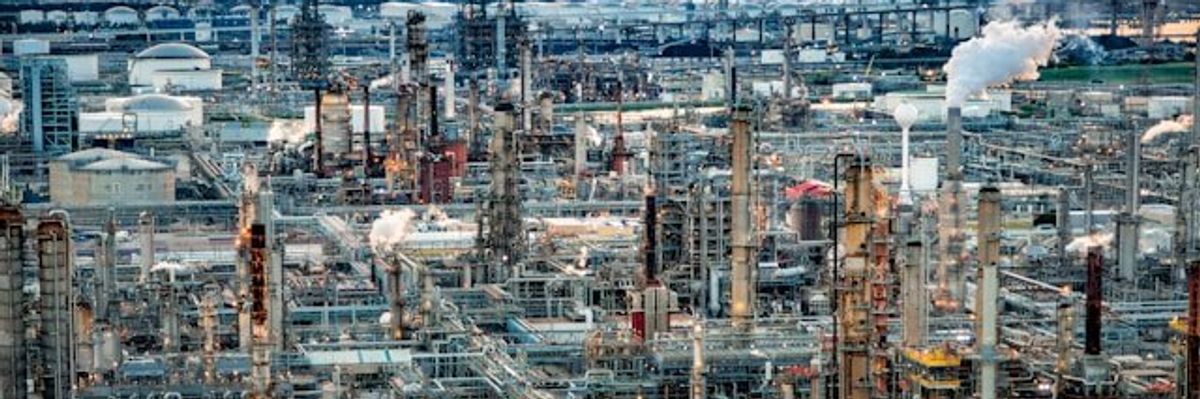 A refinery with smoke emitting from smokestacks with a city in the background.