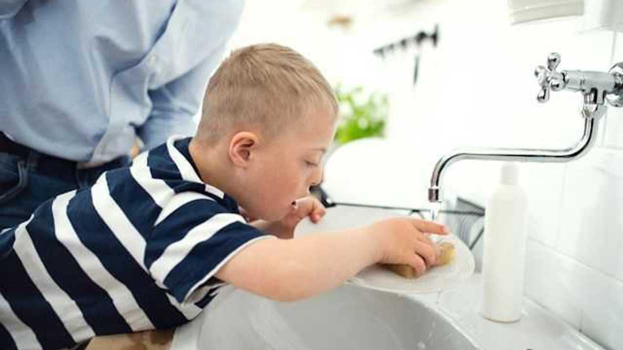 A small boy being helped by his father as he washes a dish in a kitchen sink