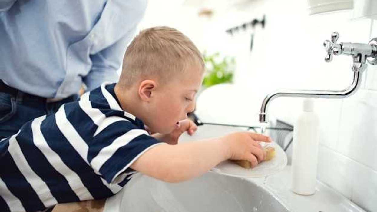 A small boy in a striped shirt washing a dish in a kitchen sink with his father supervising
