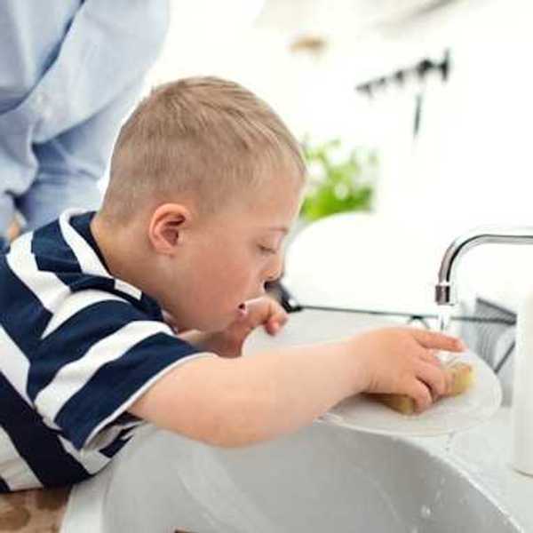 A small boy in a striped shirt washing a dish in a kitchen sink with his father supervising