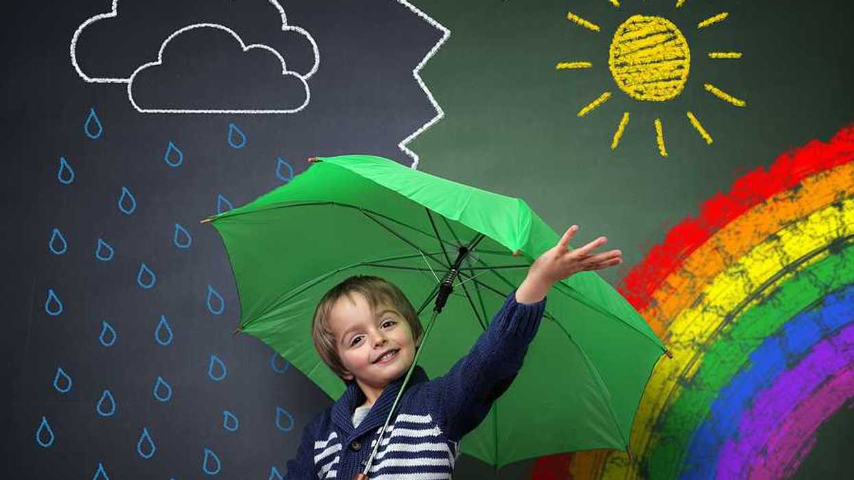 A small child under a green umbrella against a chalkboard with a rainbow and sun and rain on it