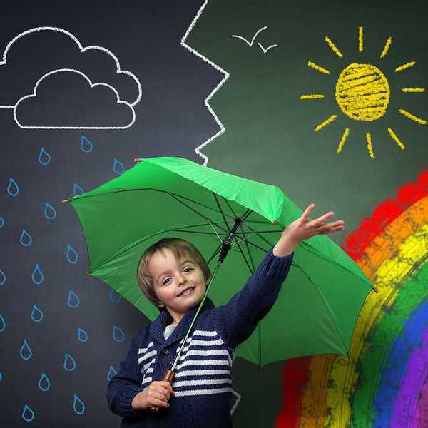 A small child under a green umbrella against a chalkboard with a rainbow and sun and rain on it