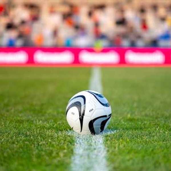 A soccer ball on green grass with people seated in bleachers in the background