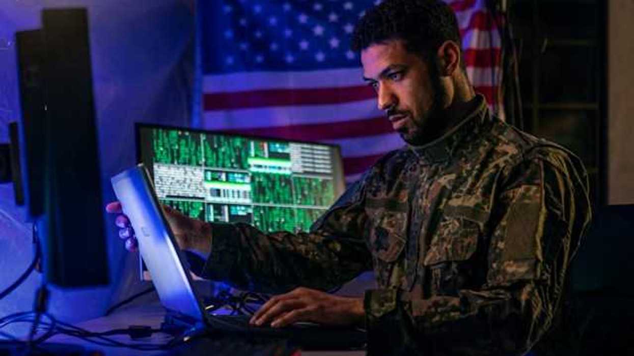 A soldier in military fatigues sitting in front of a computer with an American flag in the background