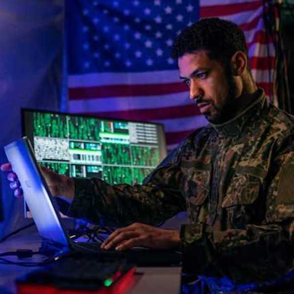 A soldier in military fatigues sitting in front of a computer with an American flag in the background
