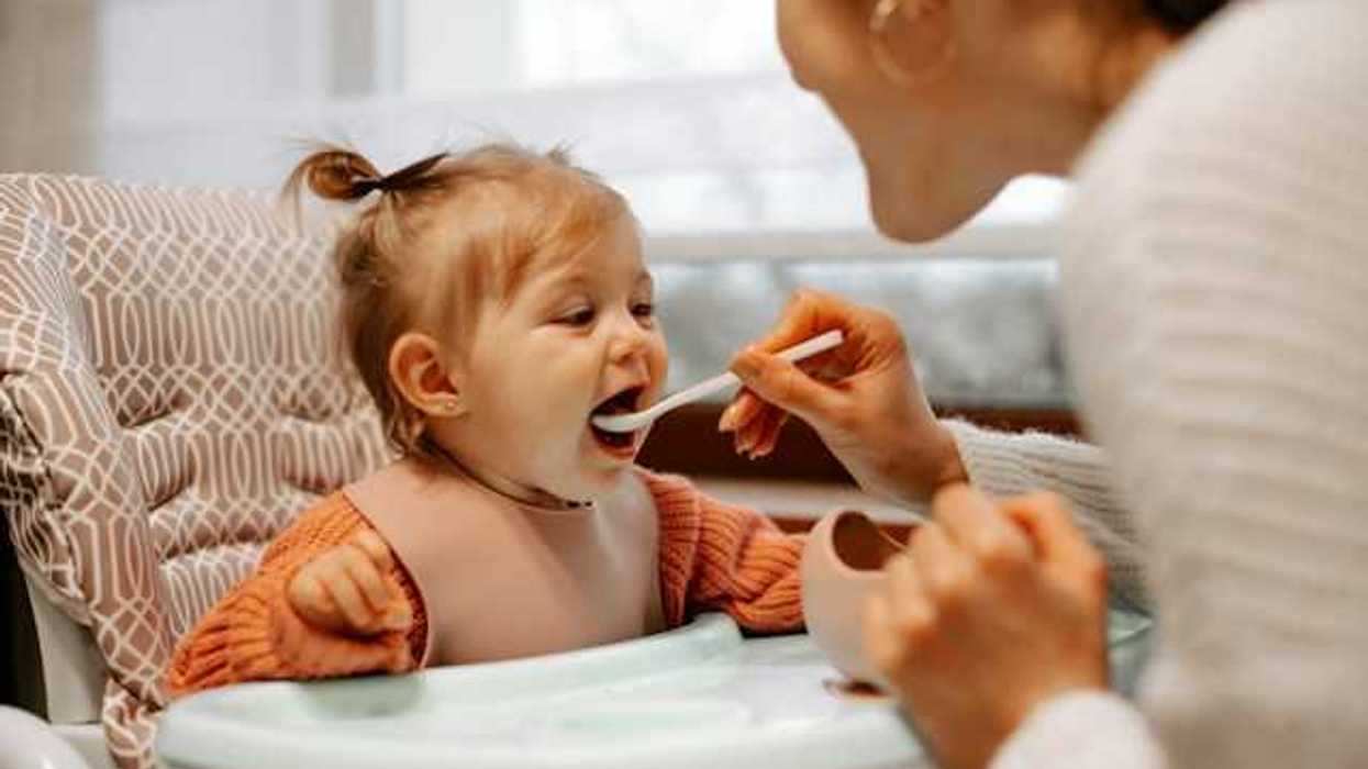 A toddler sitting in a high chair being fed by her mother