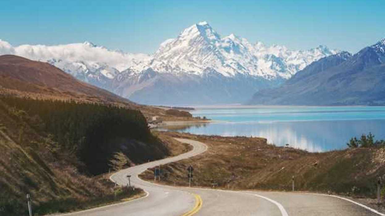 A view of a lonely highway next to an aqua lake with snow covered mountains in the background