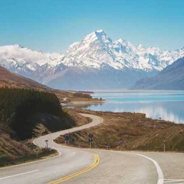 A view of a lonely highway next to an aqua lake with snow covered mountains in the background