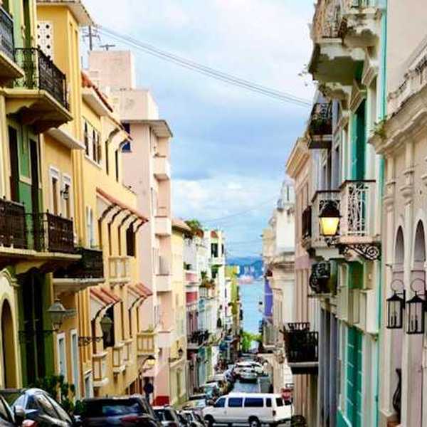 A view of a Puerto Rican street with multicolored buildings and cars and the ocean in the far distance