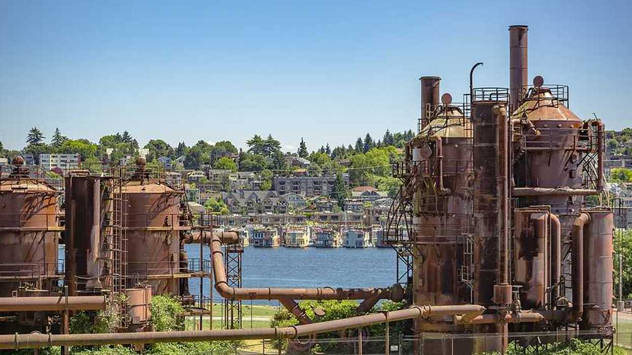 A view of rusting gas plant equipment on a green landscape with houses in the hills behind