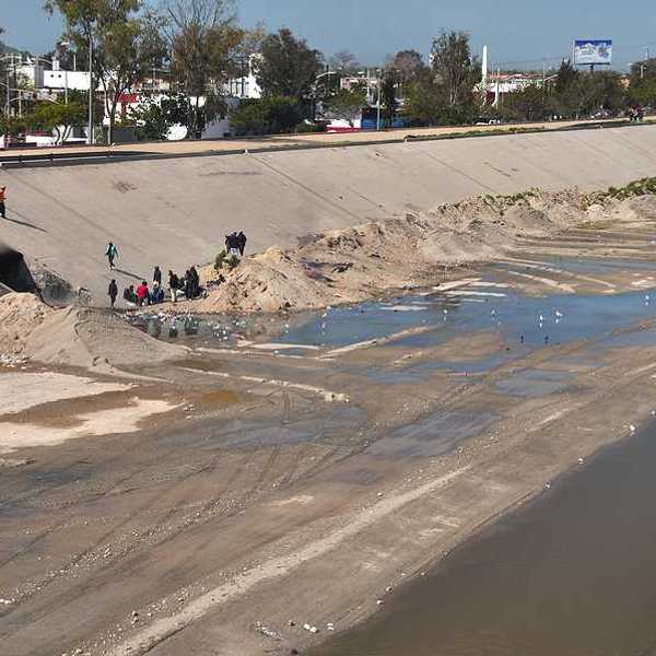 A view of the Tijuana River with water and concrete. Homeless men gather at a littered camp in the Tijuana River floodway near a pedestrian bridge.