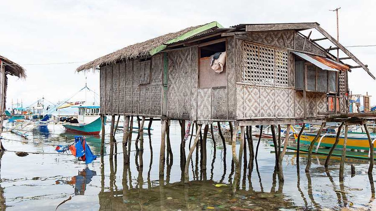 A weathered woven bamboo house on tall wooden stilts stands over shallow coastal water
