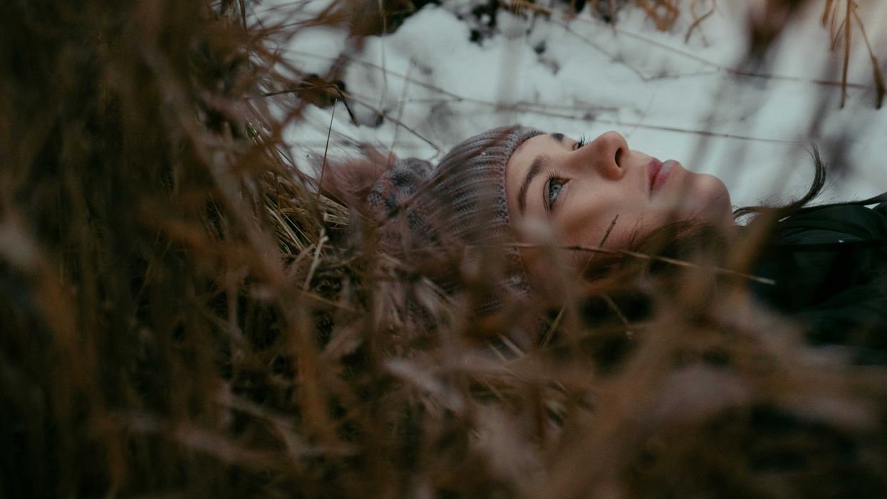 A woman in a beanie cap lays on leaves and grass looking at the sky on a cloudy day.