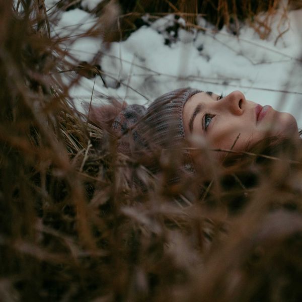 A woman in a beanie cap lays on leaves and grass looking at the sky on a cloudy day.