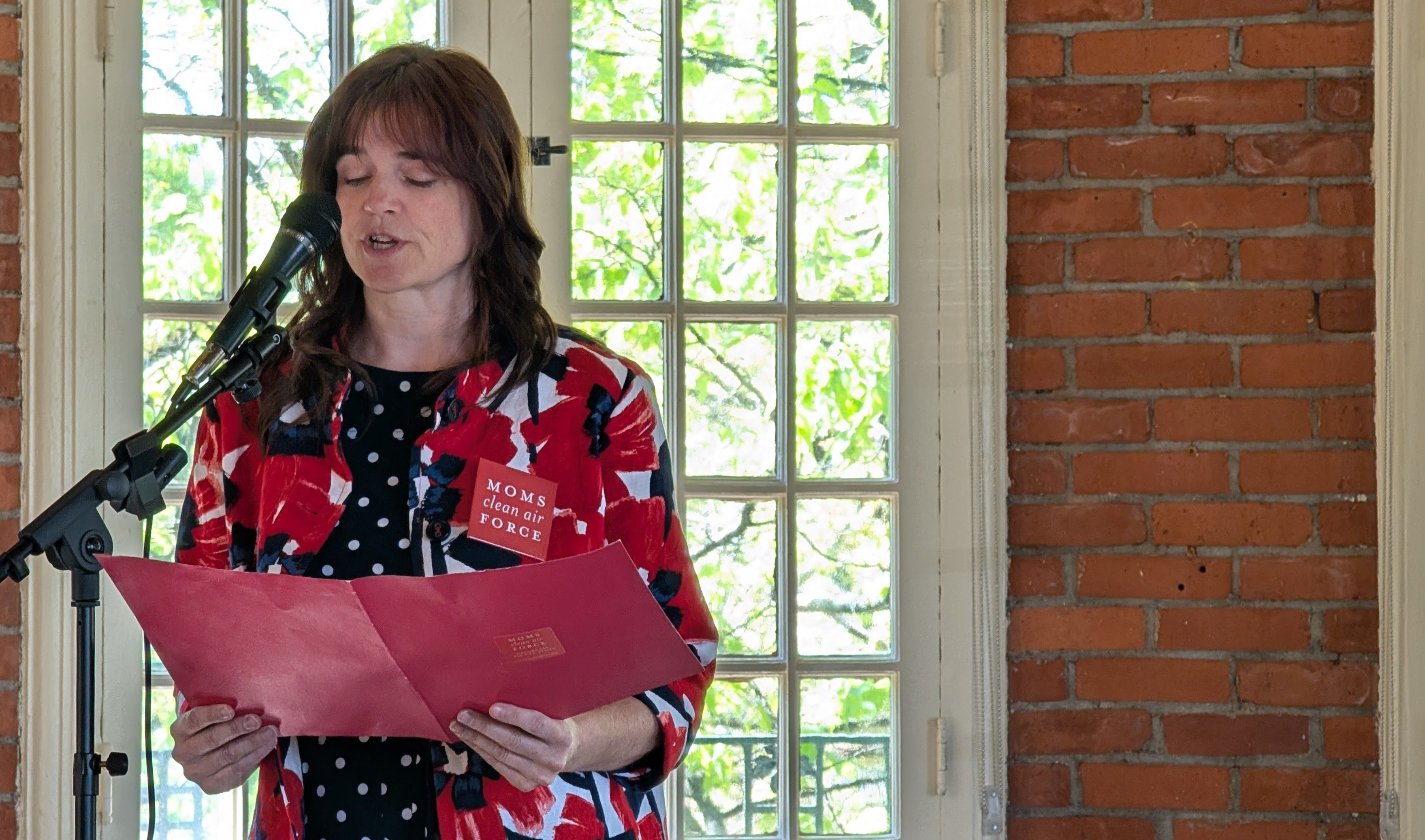 A woman in a blue polka dot dress and red floral sweater stands at a microphone in front of windows
