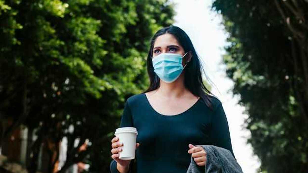 A woman in a face mask walking along a tree-lined street with a cup of coffee