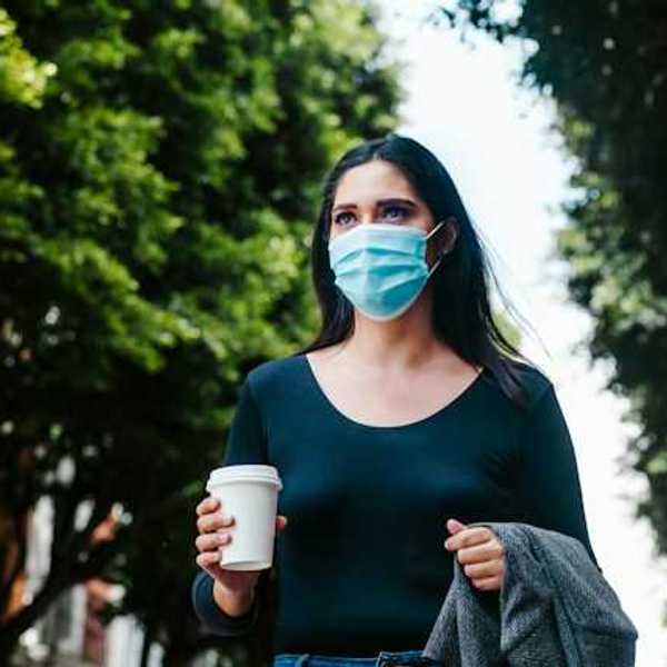 A woman in a face mask walking along a tree-lined street with a cup of coffee