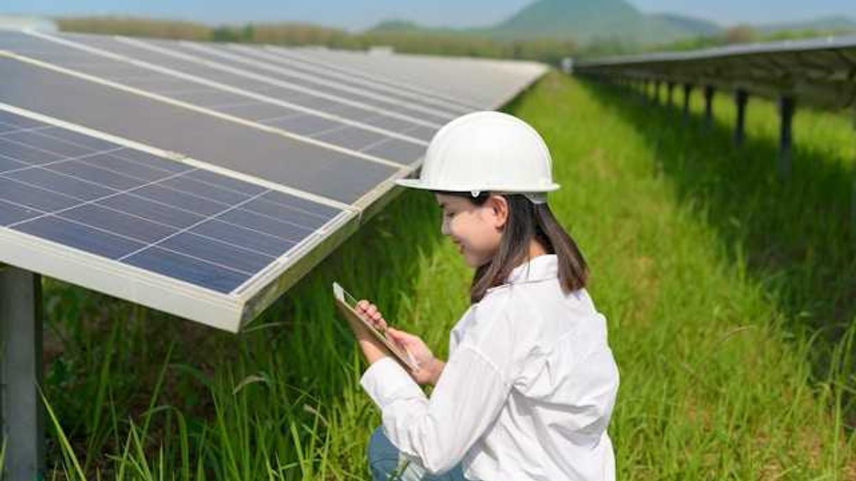 A woman in a hard hat kneels next to a solar panel in a green field holding a tablet