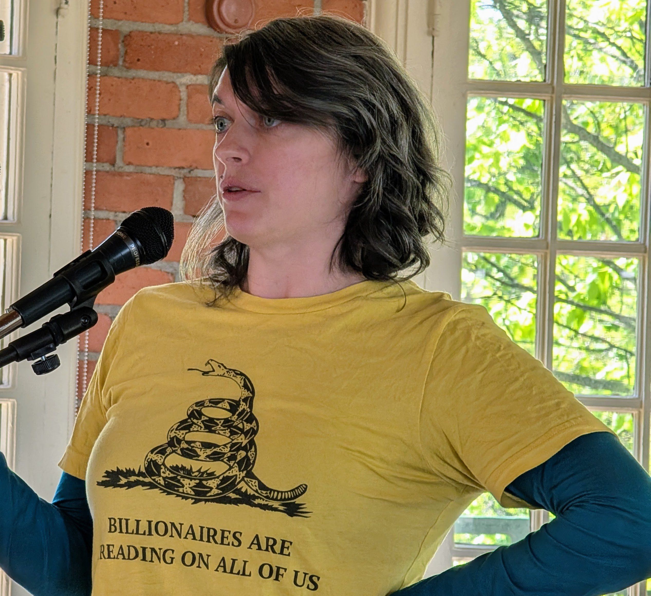 A woman in a yellow shirt stands at a microphone in front of windows
