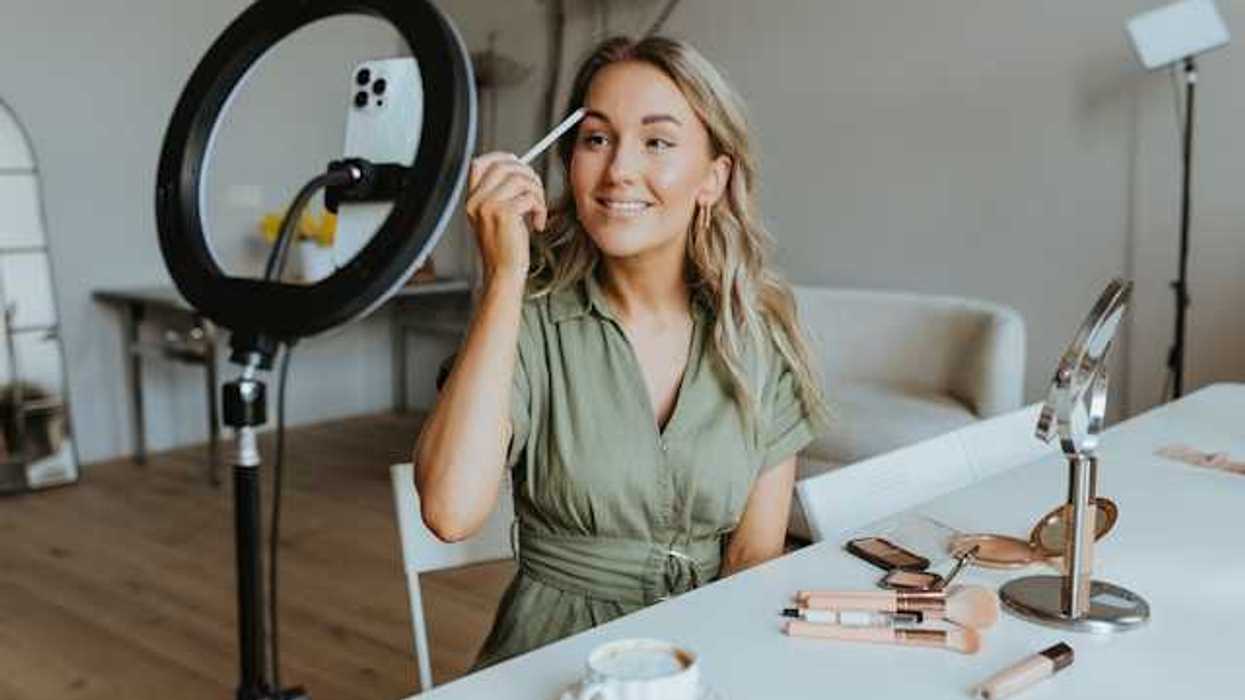 A woman in front of a cell phone and ring light putting on eyebrow makeup with cosmetics spread out on the table next to her