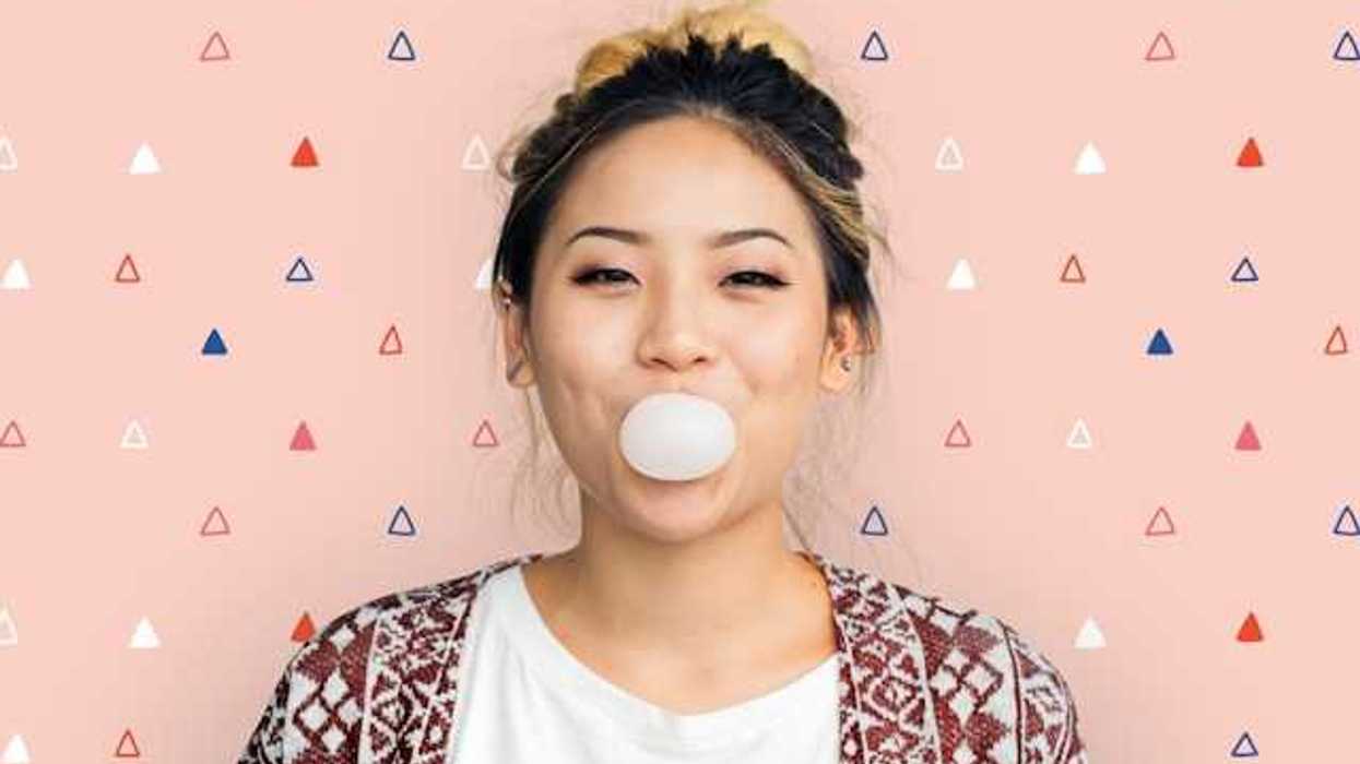 A woman standing against a pink background blowing a bubble with gum