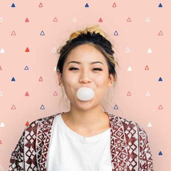 A woman standing against a pink background blowing a bubble with gum