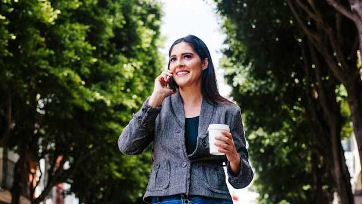 A woman walking down a tree lined street talking on the phone and carrying a takeout coffee