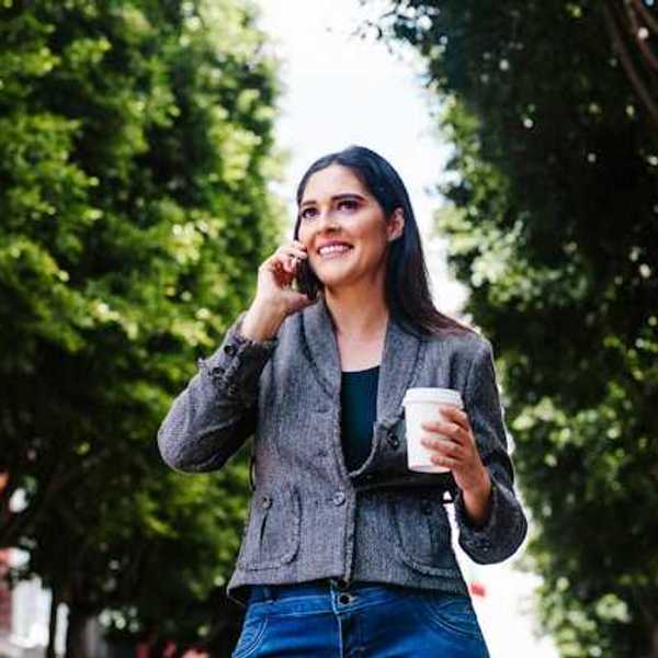 A woman walking down a tree lined street talking on the phone and carrying a takeout coffee