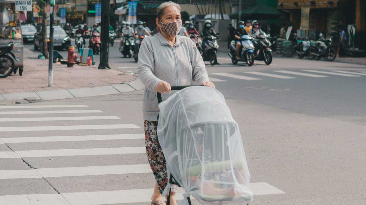 A woman wearing a face mask pushes a baby stroller across a crosswalk with traffic in background.