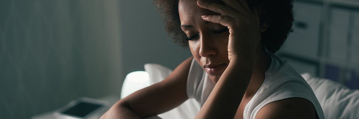 A woman wearing a tank top sitting up in bed with her hand to her forehead.