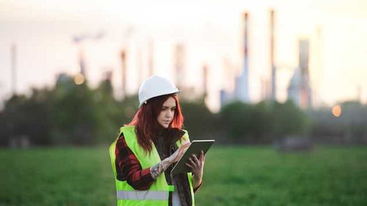 A woman with a hard hat and clip board standing in front of an oil refinery in the distance