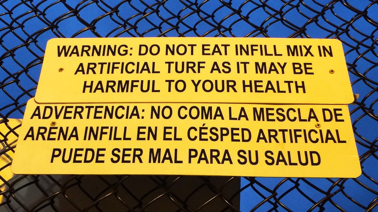 A yellow sign on a cyclone fence warns of the health dangers of artificial turf.