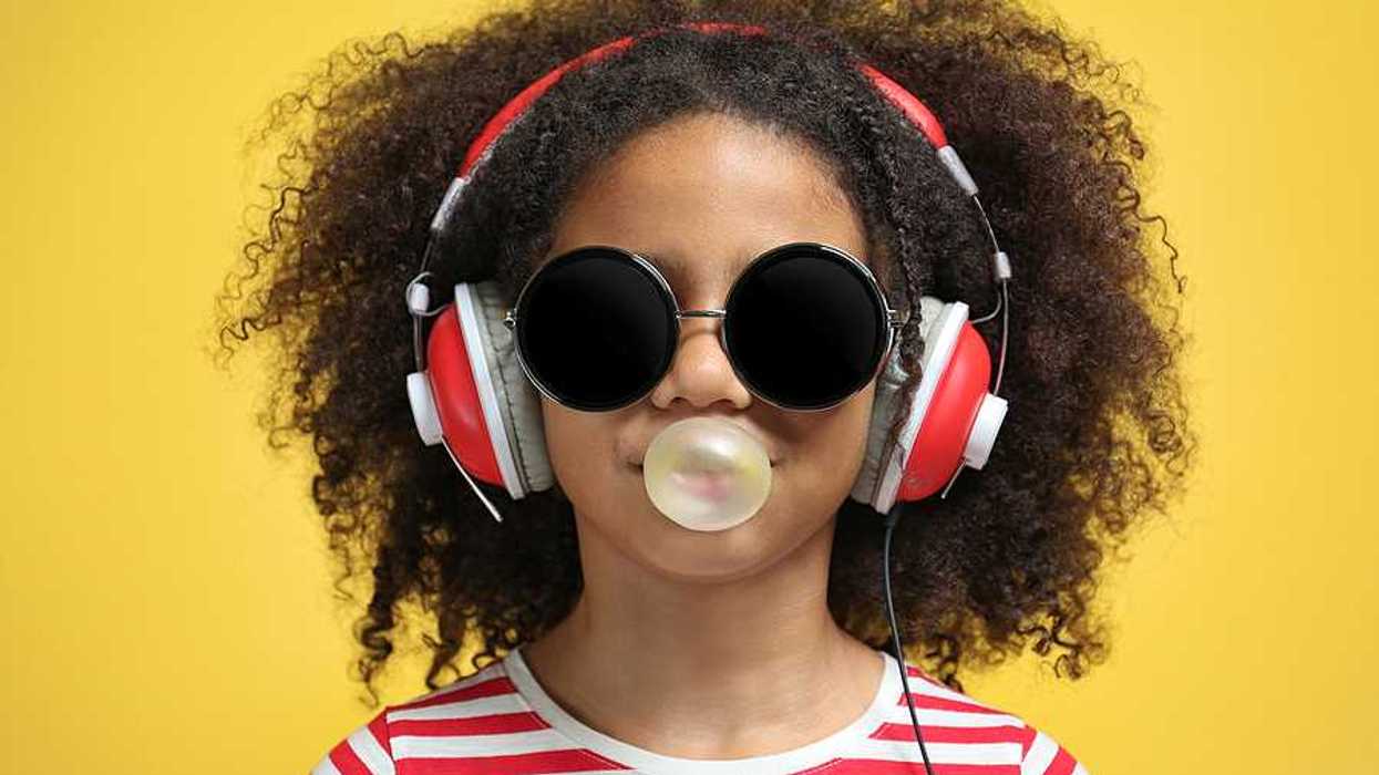 A young girl wearing headphones and a red striped shirt blowing a bubble with chewing gum
