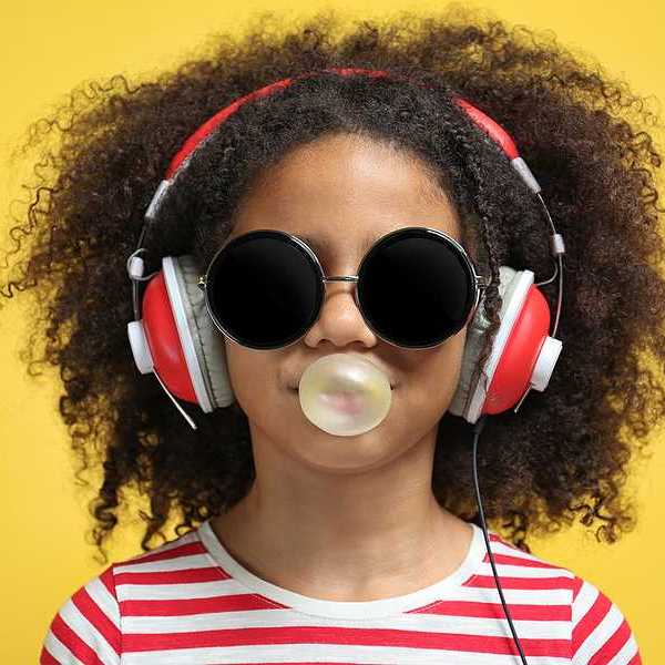 A young girl wearing headphones and a red striped shirt blowing a bubble with chewing gum