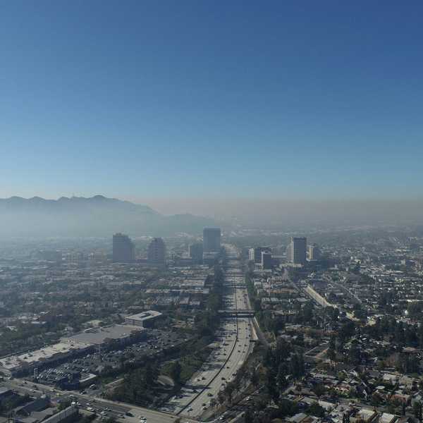 Aerial view of a city shrouded in smog criss-crossed by freeways with mountains in background.
