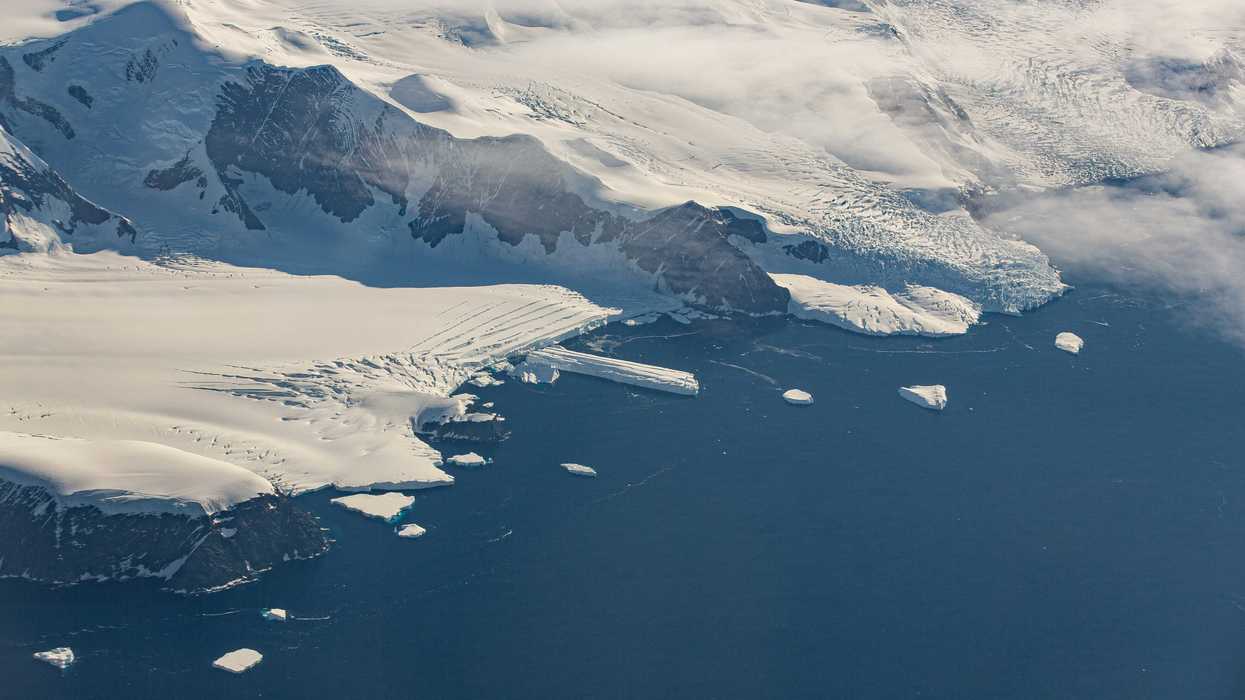 Aerial view of Antarctic coastline with glaciers and snow-covered mountains against a deep blue ocean.